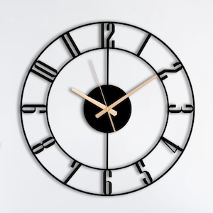 Puede incluir: Un gran reloj de pared circular con un marco de metal negro de diseño calado y números negros en negrita. El centro tiene una placa circular negra sólida con manecillas delgadas de color dorado para las horas, minutos y segundos.