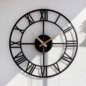 May include: A black, round wall clock with Roman numerals. The clock face is open, with the numerals and hands in black. The hands are light brown. The clock is mounted on a white brick wall.