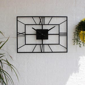 Op de afbeelding: Een zwarte rechthoekige wandklok met Romeinse cijfers en een vierkant midden. De wijzers van de klok zijn zwart en de lijst heeft een geometrisch ontwerp. De klok is bevestigd aan een witte bakstenen muur.