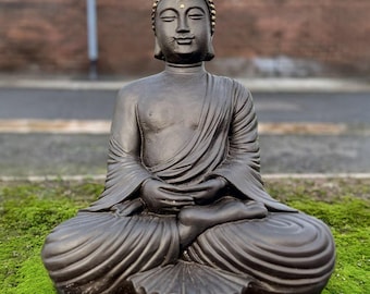 May include: A dark brown Buddha statue in a seated meditation pose. The statue has a gold-colored topknot and is set against a blurred background of brick and greenery. The statue is sitting on a bed of green moss.