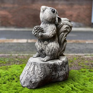 Puede incluir: Estatua de ardilla gris sosteniendo una piña, posada sobre un tocón de árbol. La ardilla mira hacia arriba. La estatua está hecha de un material similar a la piedra y está colocada sobre un lecho de musgo verde.