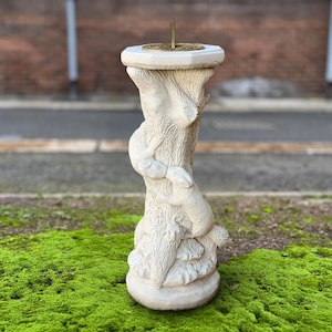 May include: A cream-coloured sundial with a textured, tree-trunk-like base and a round top. The sundial features a bronze-coloured gnomon and a decorative design on the top. The base has a sculpted design of a squirrel climbing the trunk. The sundial is outdoors on a bed of green moss.