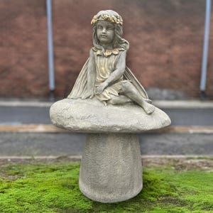 May include: A stone fairy statue with a golden flower crown and dress sits atop a mushroom-shaped pedestal. The fairy has delicate wings and is in a seated position. The statue is gray with gold accents.