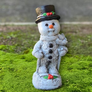 May include: A snowman figurine with a black top hat adorned with holly and berries. The snowman has a carrot nose, black button eyes, and a knitted scarf. It holds a cane and stands on a base decorated with holly. The figurine is a festive holiday decoration.