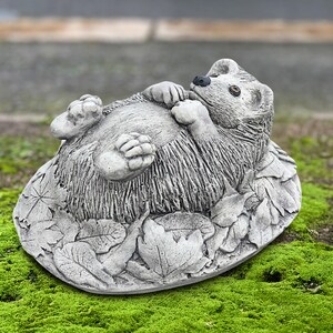 Könnte beinhalten: Eine graue Beton-Gartenstatue eines Igels, der auf dem Rücken liegt und die Pfoten in die Luft reckt. Der Igel liegt auf einem Bett aus detaillierten Blättern. Die Statue hat ein strukturiertes, stacheliges Aussehen, vor grünem Hintergrund.
