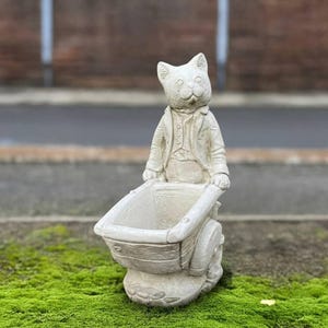 May include: A light-colored cat statue pushing a wheelbarrow. The cat is wearing a jacket and vest. The wheelbarrow has a large, open container. The statue is made of a stone-like material and is outdoors.