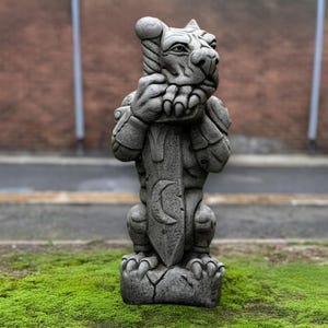 May include: A gray stone gargoyle statue with a fierce expression, holding a sword with a crescent moon design. The statue has detailed features and is set on a base, with a brick wall in the background.
