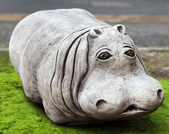 Puede incluir: Una escultura de hipopótamo de hormigón gris con rasgos faciales detallados, incluyendo orejas pequeñas y ojos expresivos. El hipopótamo descansa sobre un lecho de musgo verde vibrante, creando un contraste de texturas y colores. Decoración de jardín.