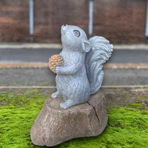 Puede incluir: Una estatua de ardilla gris sosteniendo una piña, posada sobre un tocón de árbol marrón. La ardilla mira hacia arriba. La estatua está colocada sobre un fondo de musgo verde y un fondo borroso.