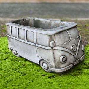 May include: A gray concrete planter shaped like a vintage Volkswagen bus. The planter has a rectangular opening for plants and features details like wheels, windows, and the VW logo. It sits on a bed of green moss.