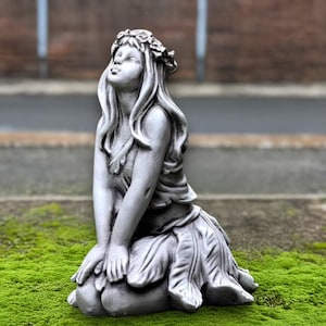May include: A gray stone garden statue of a kneeling woman with long flowing hair and a floral crown. The statue is wearing a draped dress and is set on a bed of green moss.