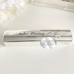 May include: Silver-plated rectangular keepsake box with the inscription "Amelia's Christenng 12.09.2025" and a small carriage design. The box is displayed on a white surface, with a framed baby photo and white flowers in the background.