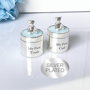 Puede incluir: Dos cajas de recuerdo plateadas, cada una con un pequeño osito de peluche en la parte superior. Una caja dice "My First Tooth" y la otra "My First Curl". Las cajas tienen una tapa azul claro y están sobre una superficie blanca.