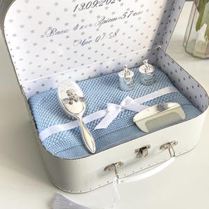 Puede incluir: Un juego de regalo para bebé en una caja con forma de maleta. La caja blanca tiene un estampado de estrellas en el interior. Contiene una manta azul, una cuchara plateada, dos tazas plateadas y un peine. Una borla blanca está unida al asa.