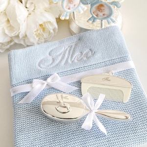 Puede incluir: Un juego de regalo para bebé con una manta de punto azul claro bordada con un nombre, un peine y un cepillo plateados. Un marco de fotos de carrusel con detalles azules y blancos está encima. Cintas y lazos blancos.