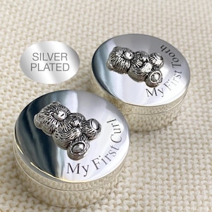 May include: Silver-plated keepsake boxes with teddy bear lid decorations. One box reads "My First Tooth" and the other reads "My First Curl". The boxes are round and sit on a cream-colored knitted surface.