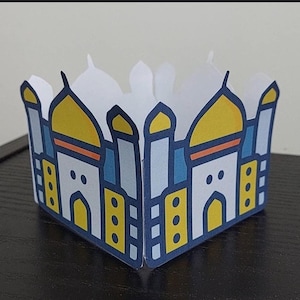May include: A white, folded paper decoration featuring a stylized illustration of a building with yellow domes, blue accents, and a navy blue border. The design is symmetrical, with the building repeated on both sides of the folded structure.