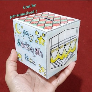 May include: A white, cube-shaped "My Sadaqah Box" with a slot on top. The box features colorful illustrations, including stars, a crescent moon, and a depiction of a religious structure. The text "Can be personalised!" is also visible.