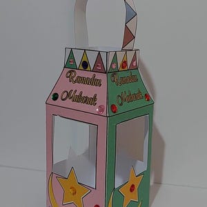 May include: A pink and green paper lantern with a handle. The lantern has a crescent moon and star design and the words "Ramadan Mubarak" on each side.