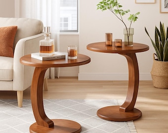 May include: Two wooden side tables with a unique curved leg design. The tables have a round top and base, and are made of a warm brown wood. One table holds a decanter and a glass, while the other has decorative glassware and a vase with greenery.