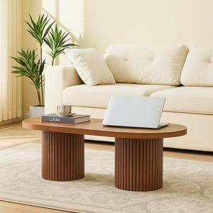 May include: A modern, oval-shaped coffee table with a dark wood finish. The table features two cylindrical, ribbed legs. A laptop, a book with the word "LOEWE" on the cover, and a decorative cup and saucer are on the table. The background includes a cream-colored sofa and a potted plant.