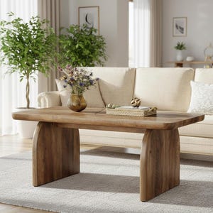May include: A rectangular wooden coffee table with rounded edges and arched legs. The table is a warm brown color and has a natural wood grain. A vase of flowers and a decorative tray sit on top of the table. The table is in front of a beige sofa.