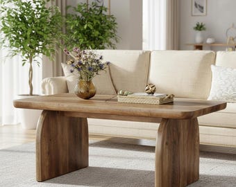 May include: A rectangular wooden coffee table with rounded edges and arched legs. The table is a warm brown color and has a natural wood grain. A vase of flowers and a decorative tray sit on top of the table. The table is in front of a beige sofa.