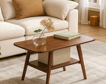 May include: A mid-century modern style coffee table with a dark wood finish. The table has a lower shelf and angled legs. A clear glass vase with dried flowers and a book are on the table. A box labeled "Medium Storage" is on the lower shelf.
