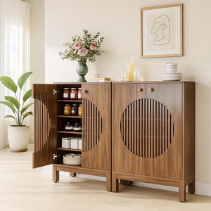 May include: A two-door wooden cabinet with a circular design on the doors. One door is open, revealing shelves with jars and containers. A vase of flowers and a framed picture are on top of the cabinet. A potted plant is to the left.