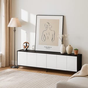 May include: A modern living room scene featuring a black and white cabinet with six doors, a framed art print, and a floor lamp. The artwork depicts a minimalist line drawing of a figure. Decorative vases and a small potted plant sit atop the cabinet.