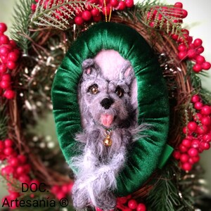 Puede incluir: Un adorno de perro de fieltro gris y blanco con un fondo de terciopelo verde. El adorno tiene forma ovalada y tiene una campana dorada colgando del collar del perro. El adorno está colgado de una corona de Navidad con bayas rojas.