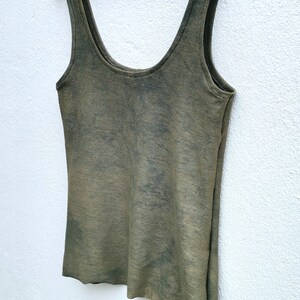 May include: A green sleeveless tank top with a tie-dye pattern. The tank top is hanging on a white hanger against a white wall.
