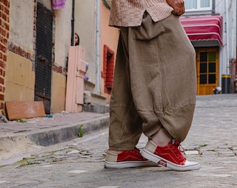 Könnte beinhalten: Eine Person in einem hellbraunen Strickhemd und einer lockeren braunen Hose steht auf einer Kopfsteinpflasterstraße. Rote Turnschuhe mit weißen Sohlen und Akzenten sind zu sehen. Im Hintergrund sind Gebäude und Wäsche zu sehen.