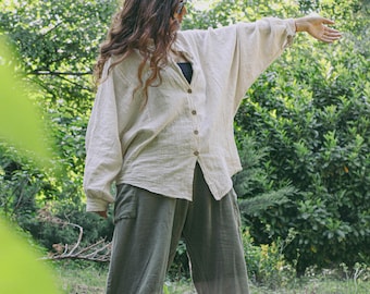 May include: A light beige button-down shirt with wide sleeves and olive green pants. The outfit is paired with white sneakers. The person is standing outdoors in a natural setting, surrounded by greenery.