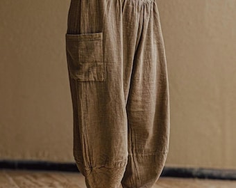 May include: Brown harem pants with a wide, textured waistband and a single pocket. The pants have a relaxed fit and are paired with orange sneakers with white laces and soles. The person is wearing a white top.