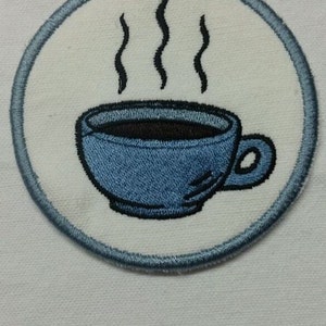 May include: A blue and white embroidered iron-on patch with a steaming cup of coffee. The patch is circular and has a light blue border.