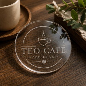 Pode incluir: Um porta-copos circular de acrílico transparente com um design gravado que inclui um ícone de chávena de café, o texto TEO CAFE COFFEE CO. e um gráfico de grão de café centrado dentro de uma borda circular na superfície transparente.