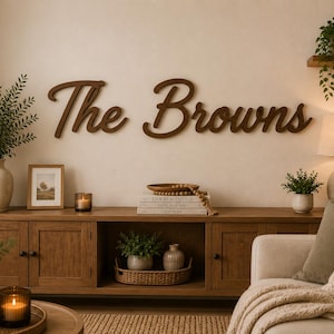 Può includere: Una scritta da parete in legno marrone scuro che riporta il testo The Browns in un carattere corsivo. La scritta è montata su una parete di colore chiaro sopra un mobile multimediale in legno, decorato con varie piante, candele e libri.