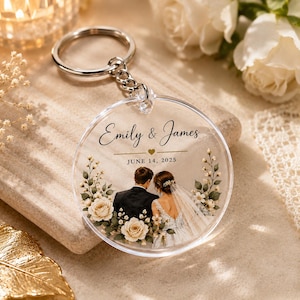 Puede incluir: Un llavero circular de acrílico transparente con una ilustración impresa de una pareja con ropa de boda. El texto dice Emily & James y JUNE 14, 2025, separados por un pequeño corazón. El diseño incluye una corona de rosas color crema.