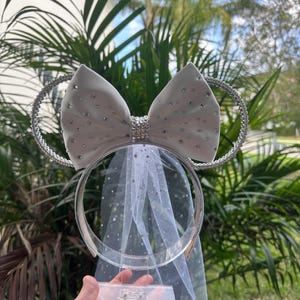 May include: White mouse ears headband with a large white satin bow decorated with silver rhinestones. The headband has a white tulle veil attached.