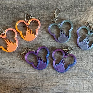 May include: Six keychains in the shape of Mickey Mouse's head, each with a castle silhouette inside. The keychains have a gradient color scheme, ranging from orange to purple, and are attached to silver-colored clasps.