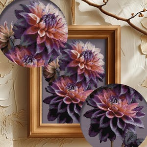 May include: A framed floral artwork featuring a detailed cross-stitch design of dahlias in shades of purple, peach, and green. The artwork is set in a wooden frame, with two circular close-ups of the flowers. The background is a neutral gray.