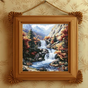 Könnte beinhalten: Gerahmte Kreuzstich-Landschaft mit einem Wasserfall, der in einen blauen Fluss stürzt, umgeben von herbstlich gefärbten Bäumen und Bergen. Der Holzrahmen hat dekorative Eckakzente und wird von einem Seil gehalten.