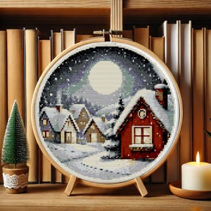 May include: A cross-stitch embroidery hoop with a winter scene of a snowy village under a full moon. The houses are red, brown, and white with snow-covered roofs. The background is a dark blue sky with white snowflakes.