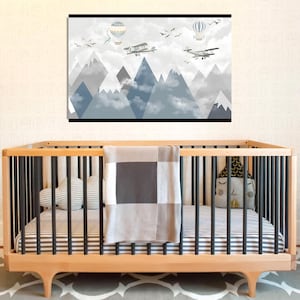 May include: A nursery scene featuring a wooden crib with a striped mattress, a gray and white blanket, and decorative pillows. Above the crib is a framed wall art depicting mountains, clouds, airplanes, hot air balloons, and birds in shades of gray and blue.