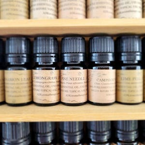 May include: A close-up of a wooden shelf with rows of small, dark brown glass bottles filled with essential oils. The bottles have labels with the names of the oils, including "Lemongrass", "Pine Needle", and "Camphor".
