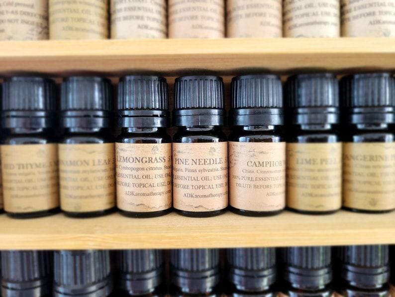 May include: A close-up of a wooden shelf with rows of small, dark brown glass bottles filled with essential oils. The bottles have labels with the names of the oils, including "Lemongrass", "Pine Needle", and "Camphor".