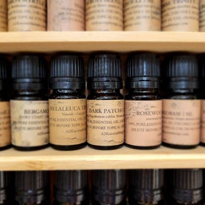 May include: A shelf of small, dark brown glass bottles with labels. The labels are white with black text and list the names of essential oils, including "Bergamot", "Melaleuca Tea Tree", "Dark Patchouli", "Rosewood", and "Mandarin".