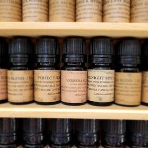 May include: A close-up of a shelf with small glass bottles filled with essential oils. The bottles have black caps and labels with text that includes the names "Unicorn Blend", "Perfect Peach", "Verbena Blend", "Midnight Special", and "Terra Blend".