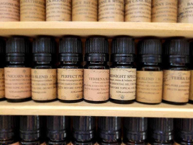 May include: A close-up of a shelf with small glass bottles filled with essential oils. The bottles have black caps and labels with text that includes the names "Unicorn Blend", "Perfect Peach", "Verbena Blend", "Midnight Special", and "Terra Blend".